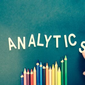 Colored pencils and letters spelling 'ANALYTICS' on a textured background with a hand.